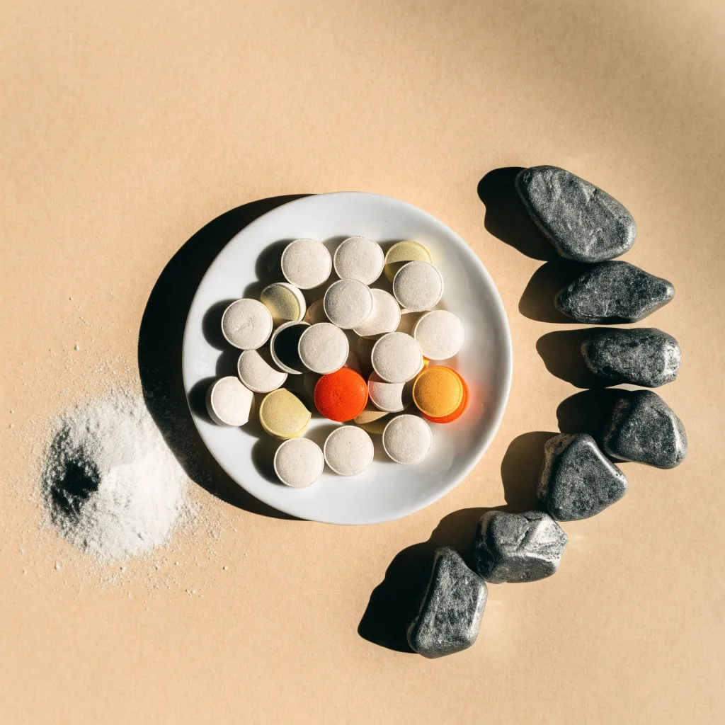 Tabletas multivitamínicas sin marca en un plato llano junto a granos de magnesio y piedras grises de zinc